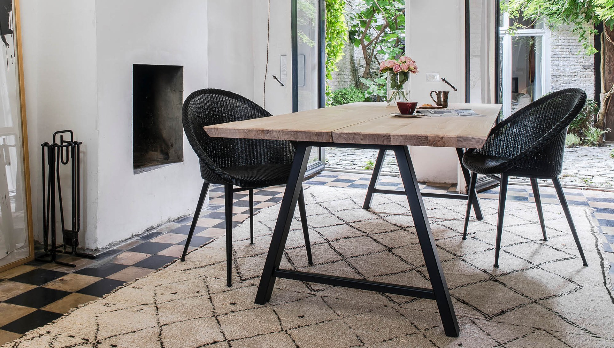 Dining room featuring a Vincent Sheppard Albert dining table with a wooden top and black metal legs, paired with black Jack dining chairs. The room opens to a garden courtyard and has a chequered floor with a patterned rug.