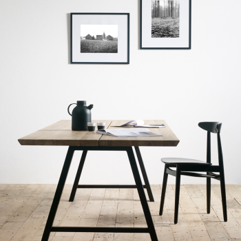 Eethoek met een Vincent Sheppard Albert A Base eettafel met houten blad en zwarte metalen poten, gecombineerd met een zwarte Teo eetkamerstoel. De ruimte bevat twee zwart-wit ingelijste foto's aan een witte muur.