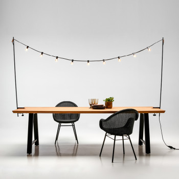 Vincent Sheppard Matteo dining table with an untreated teak top and black A-frame legs, accompanied by Edgard dining chairs with a steel base in black, under a string of lights from the "Light My Table" collection.