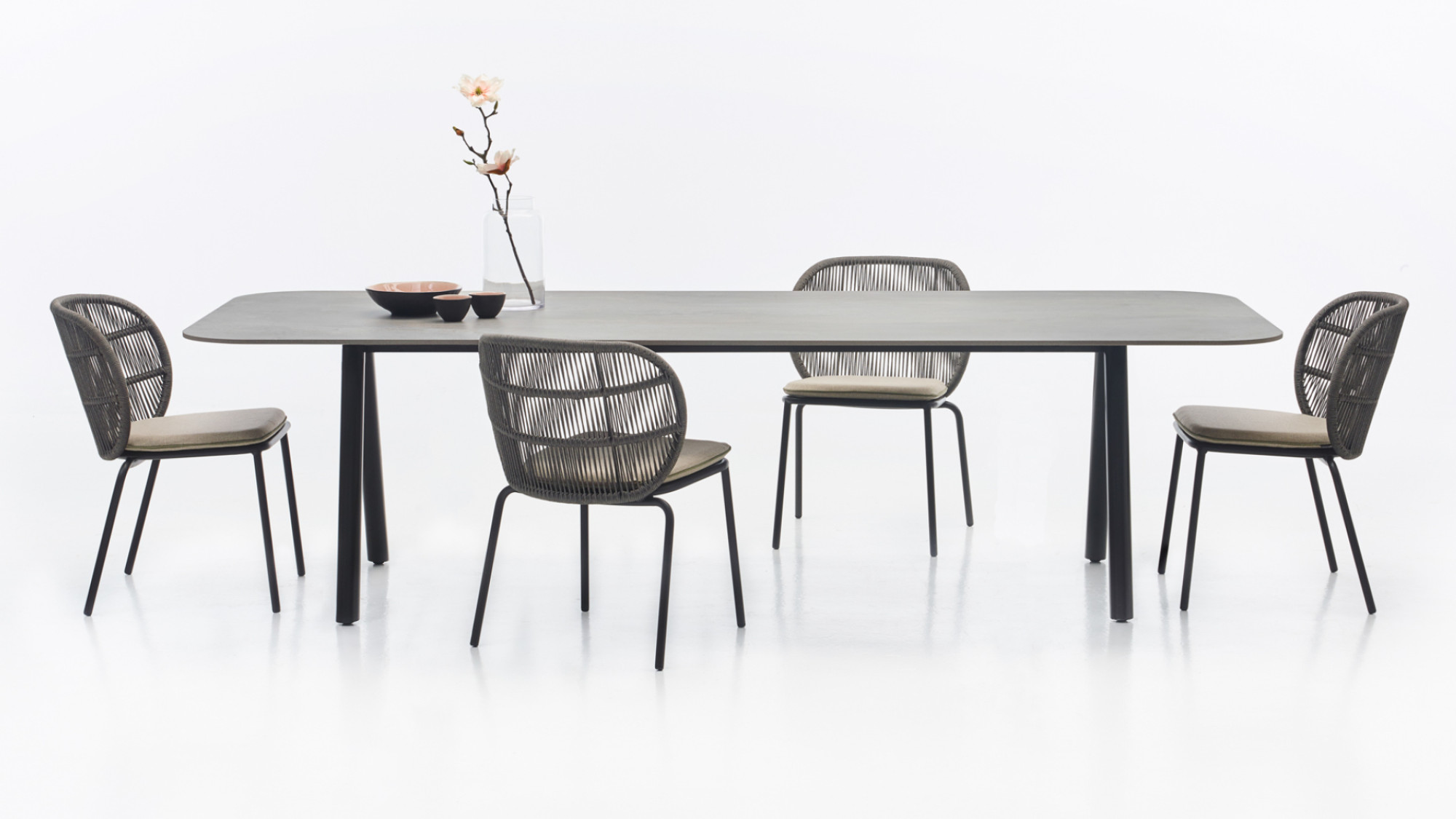 Vincent Sheppard Kodo dining table with flint-coloured top and fossil grey legs, accompanied by four Kodo dining chairs in fossil grey, featuring rope backs and cushioned seats. The minimalist table setting includes a vase with a single flower and ceramic bowls.