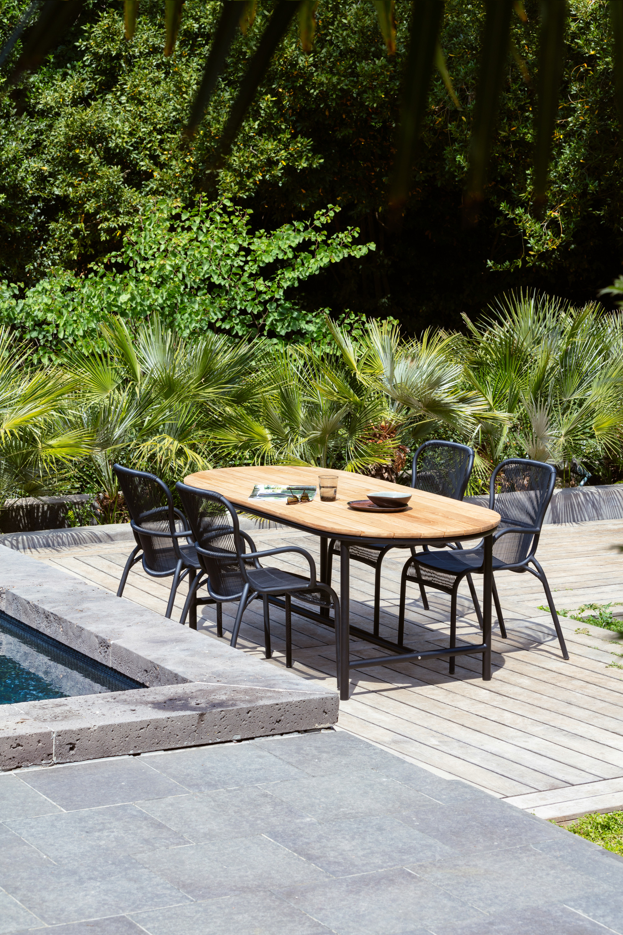 Poolside dining area featuring a Vincent Sheppard Wicked dining table with a natural wood top and matching Loop dining chairs, surrounded by lush greenery.