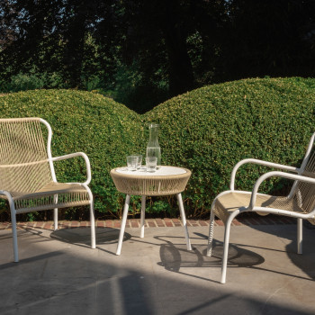 Buitenopstelling met twee Vincent Sheppard Loop loungestoelen met gevlochten zitting en een bijpassende bijzettafel, op een terras met weelderig groen op de achtergrond.