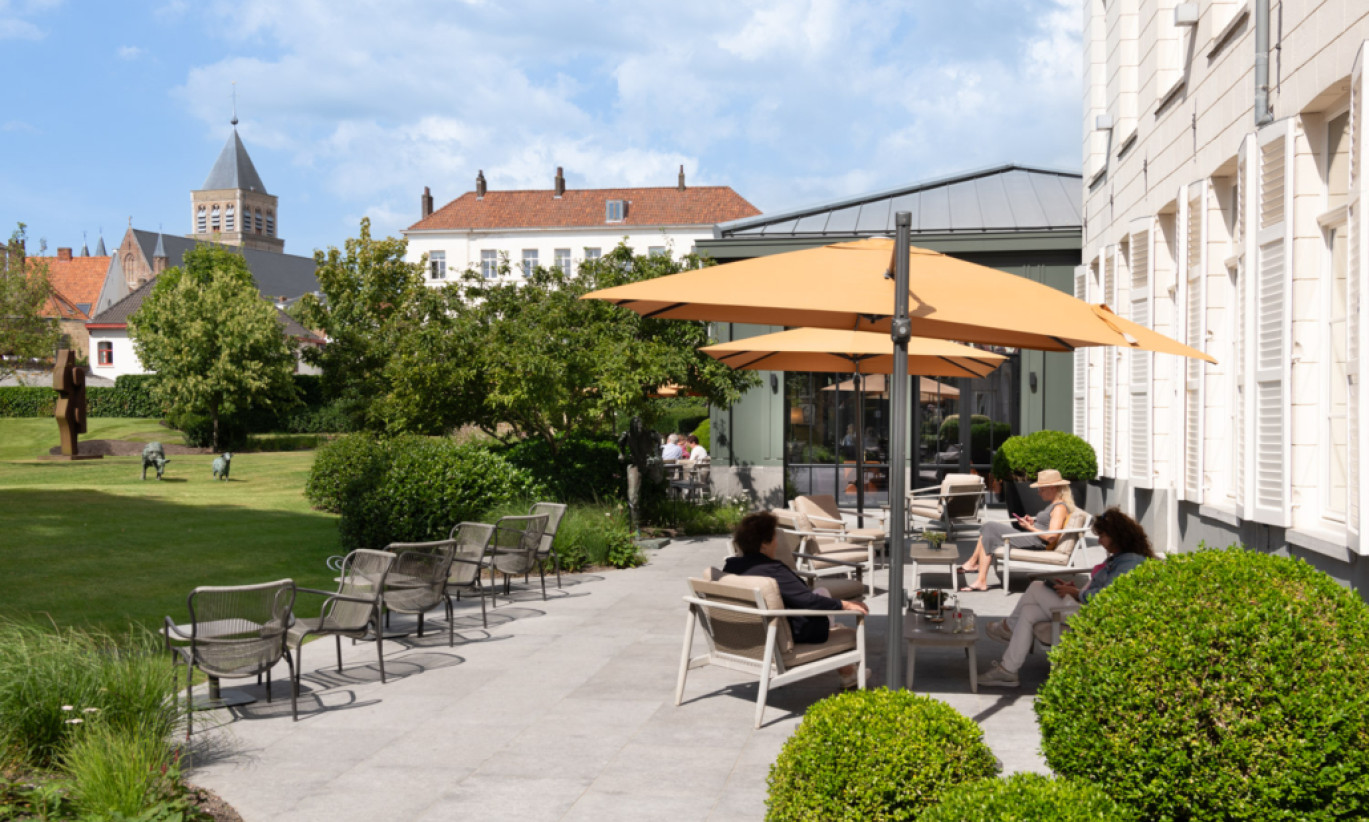 Espace de détente extérieur au Dukes Palace Brugge avec des chaises longues Loop, des chaises longues David et des canapés David sous des parasols jaunes. Un jardin avec des sculptures et des bâtiments historiques en arrière-plan.