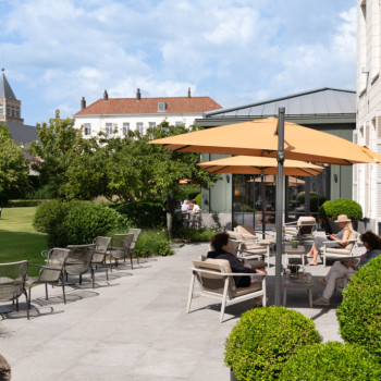 Espace de détente extérieur au Dukes Palace Brugge avec des chaises longues Loop, des chaises longues David et des canapés David sous des parasols jaunes. Un jardin avec des sculptures et des bâtiments historiques en arrière-plan.