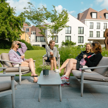 Vier Frauen sitzen und plaudern auf David Lounge-Sesseln und -Sofas im Garten des Duke's Palace mit modernen Gebäuden im Hintergrund.