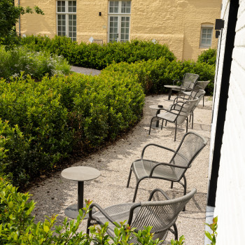 Espace de sièges dans le jardin au Duke's Palace à Brugge avec des chaises longues Loop et des petites tables rondes, entouré de verdure et face à un bâtiment jaune avec toit en tuiles rouges.