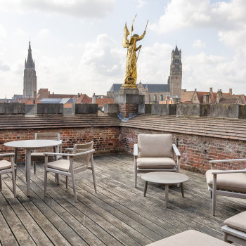 Dakterras bij Duke's Palace in Brugge met David eetkamerstoelen, David loungestoelen en ronde tafels, met een gouden standbeeld en uitzicht op de skyline van de stad.