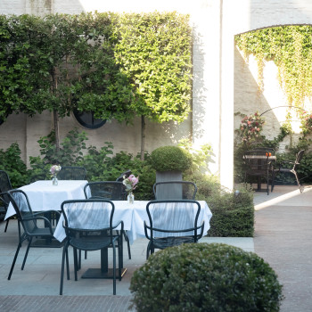 Buiteneetruimte bij Cobergher Hotel in Kortrijk met tafels bedekt met witte tafelkleden, Loop eetstoelen en omliggend groen.