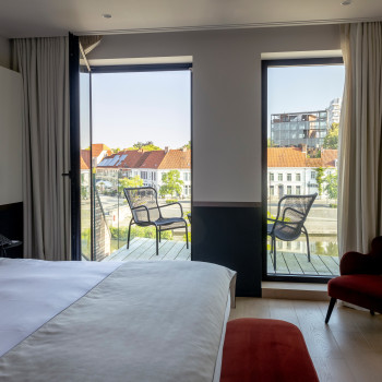 Hotelzimmer im Cobergher Hotel in Kortrijk mit einem Kingsize-Bett, einem roten Samtsessel und einer Terrasse mit Loop-Loungesesseln mit Blick auf die Stadt.