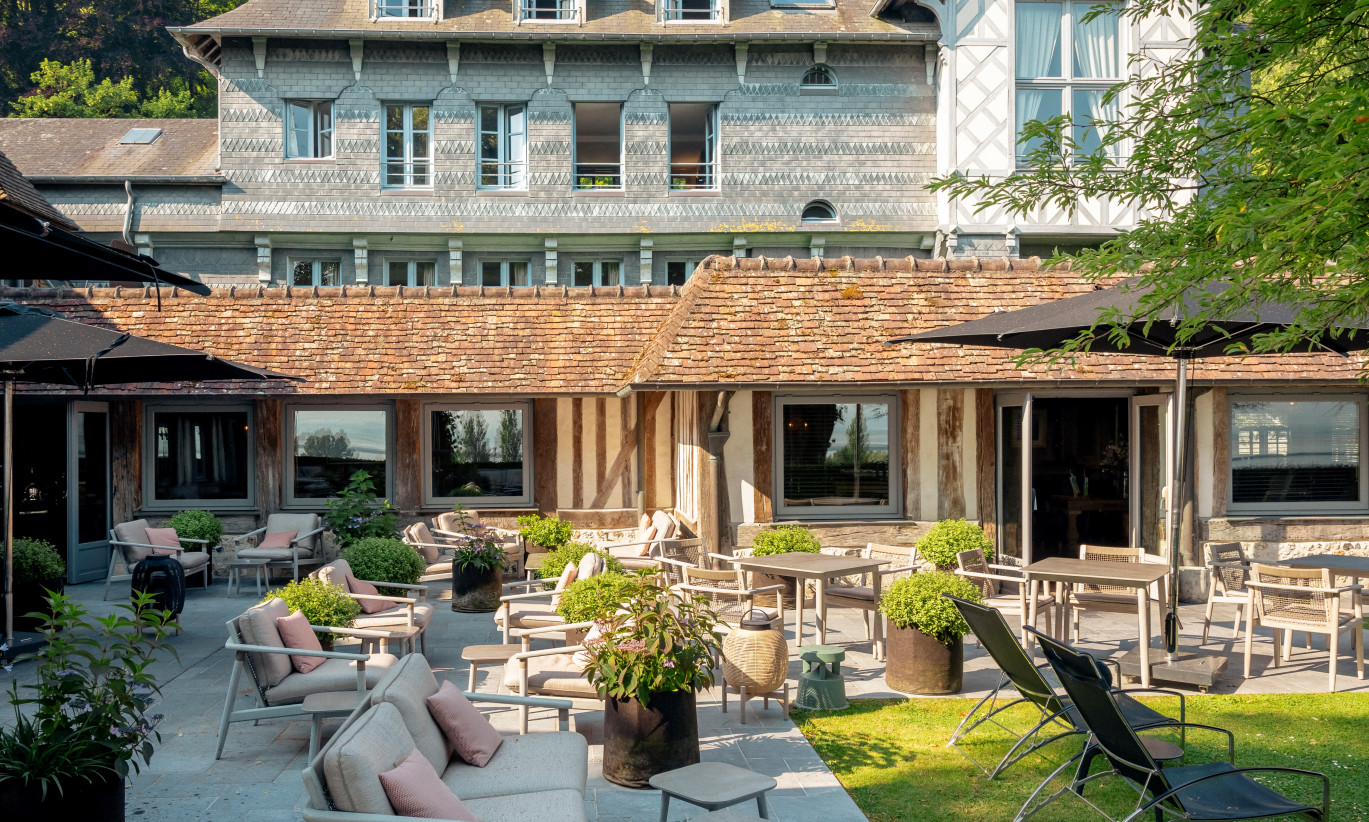  Buiten zitgedeelte bij La Ferme Saint-Siméon met David loungesofa's, David lounge stoelen en David bijzettafels, omringd door groen en in de schaduw van parasols.