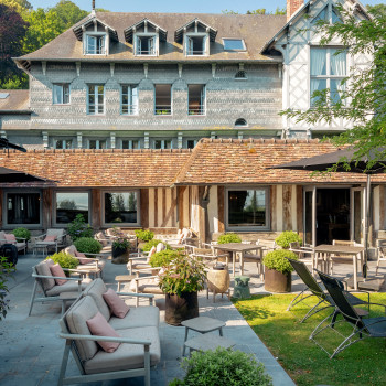  Buiten zitgedeelte bij La Ferme Saint-Siméon met David loungesofa's, David lounge stoelen en David bijzettafels, omringd door groen en in de schaduw van parasols.