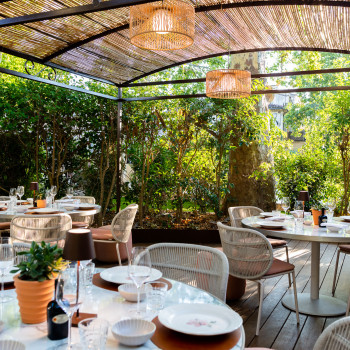 Buiten eetgedeelte bij La Bastide Bourrelly met ronde tafels gedekt met borden en glazen, omringd door Kodo eetstoelen onder een geweven overkapping, en weelderig groen op de achtergrond.