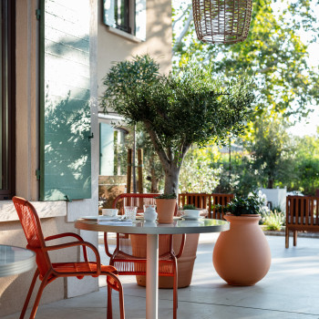 Buiten zitgedeelte bij La Bastide Bourrelly met een kleine ronde tafel gedekt voor twee, omringd door terracotta Loop eetstoelen, potplanten en een grote olijfboom onder een gevlochten hanglamp.