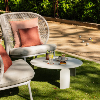 Sitzbereich im Freien bei La Bastide Bourrelly mit Kodo Cocoon-Stühlen in Dune White, rostfarbenen Kissen, einem niedrigen runden Tisch mit Getränken und Snacks und einem Pétanque-Platz im Hintergrund.