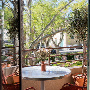 Coin salon sur le balcon à La Bastide Bourrelly avec une table ronde en marbre, des chaises de salle à manger Loop en terracotta, un vase de fleurs séchées, et une vue sur les arbres et le jardin en contrebas.