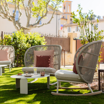 Gartensitzbereich bei La Bastide Bourrelly mit Kodo-Schaukelstühlen in Dune White und rostfarbenen Kissen, einem niedrigen runden Tisch mit Getränken und Snacks, umgeben von Grün und Blick auf nahegelegene Gebäude.