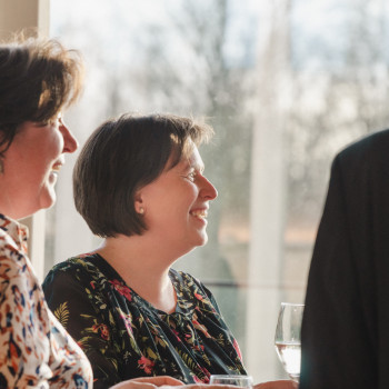 Two smiling colleagues from Vincent Sheppard enjoying a conversation indoors, with sunlight streaming through a large window in the background.