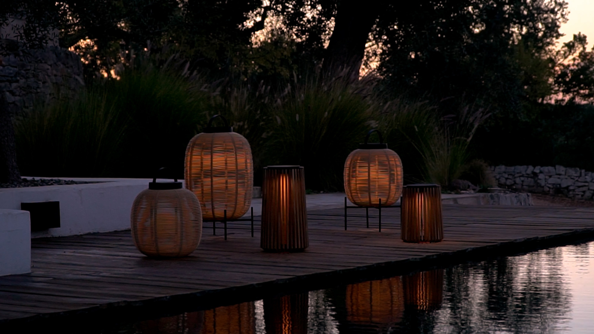 Cadre extérieur au crépuscule avec les lanternes Tika et Maya de Vincent Sheppard diffusant une lumière chaude sur une terrasse en bois, reflétées dans un point d'eau à proximité, entourées de verdure.