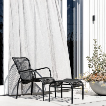 Fauteuil lounge Vincent Sheppard Loop en noir avec repose-pieds Loop assorti sur une terrasse en bois blanc, à côté d’un grand pot avec des plantes vertes et jaunes