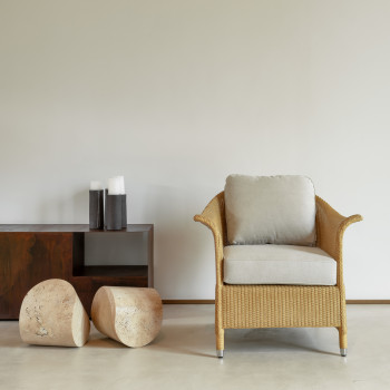  Fauteuil Victor de Vincent Sheppard avec un coussin beige, placé à côté d'une table d'appoint en bois et de deux tabourets en pierre sur un sol minimaliste.