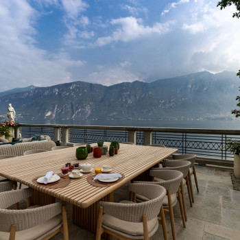 Outdoor dining area at Breakwater Bellagio with a large wooden table, Mona chairs, and a view of Lake Como and the surrounding mountains. The table is set with breakfast items and colourful vases, with a lounge area in the background.