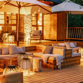 Coin salon extérieur confortable au camp Bushtops, avec des fauteuils lounge Leo et des tables basses modulaires, éclairé par des lanternes Tika. La terrasse en bois est aménagée avec des coussins et des plaids, créant une atmosphère chaleureuse et accueillante sous de grands parasols.