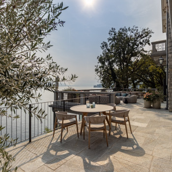 Terrace at Breakwater Bellagio with a dining setup featuring Mona chairs and a round wooden table, overlooking Lake Como with surrounding olive trees and lounge areas.