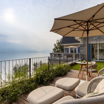 Lakeside terrace at Breakwater Bellagio with Kodo Cocoon chairs and footrests under a large parasol, offering views of Lake Como and surrounding mountains. The stone house features blue shutters and a balcony.