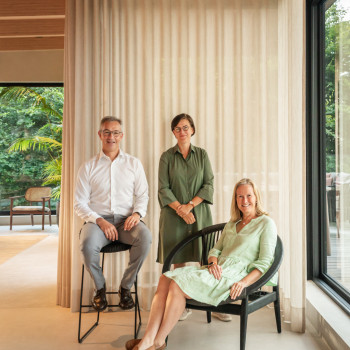 Vincent Sheppard management team posing indoors with modern furniture, including a black armchair and a barstool, standing in front of light beige curtains with a view of greenery outside.