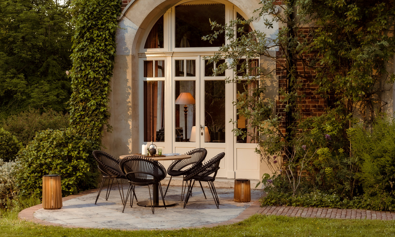 Terrasse extérieure d'une maison de campagne avec des chaises Roxy, une table bistro Ronda et des lampes Maya. Le cadre présente des murs recouverts de lierre et une vue sur le jardin.