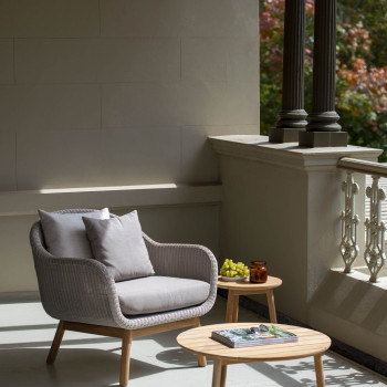 Anton lounge chair in Old Lace with a wicker seat and teak frame, paired with the Anton coffee table and side table, both in teak, by Vincent Sheppard. A small plate of grapes and a candle sit on the side table, with a magazine on the coffee table, on a shaded balcony with decorative railing and columns.