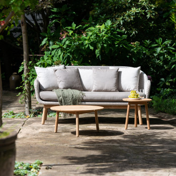 Garden seating area with an Anton lounge sofa and two Anton coffee tables, surrounded by greenery. A green throw and a plate of grapes are placed on the furniture.