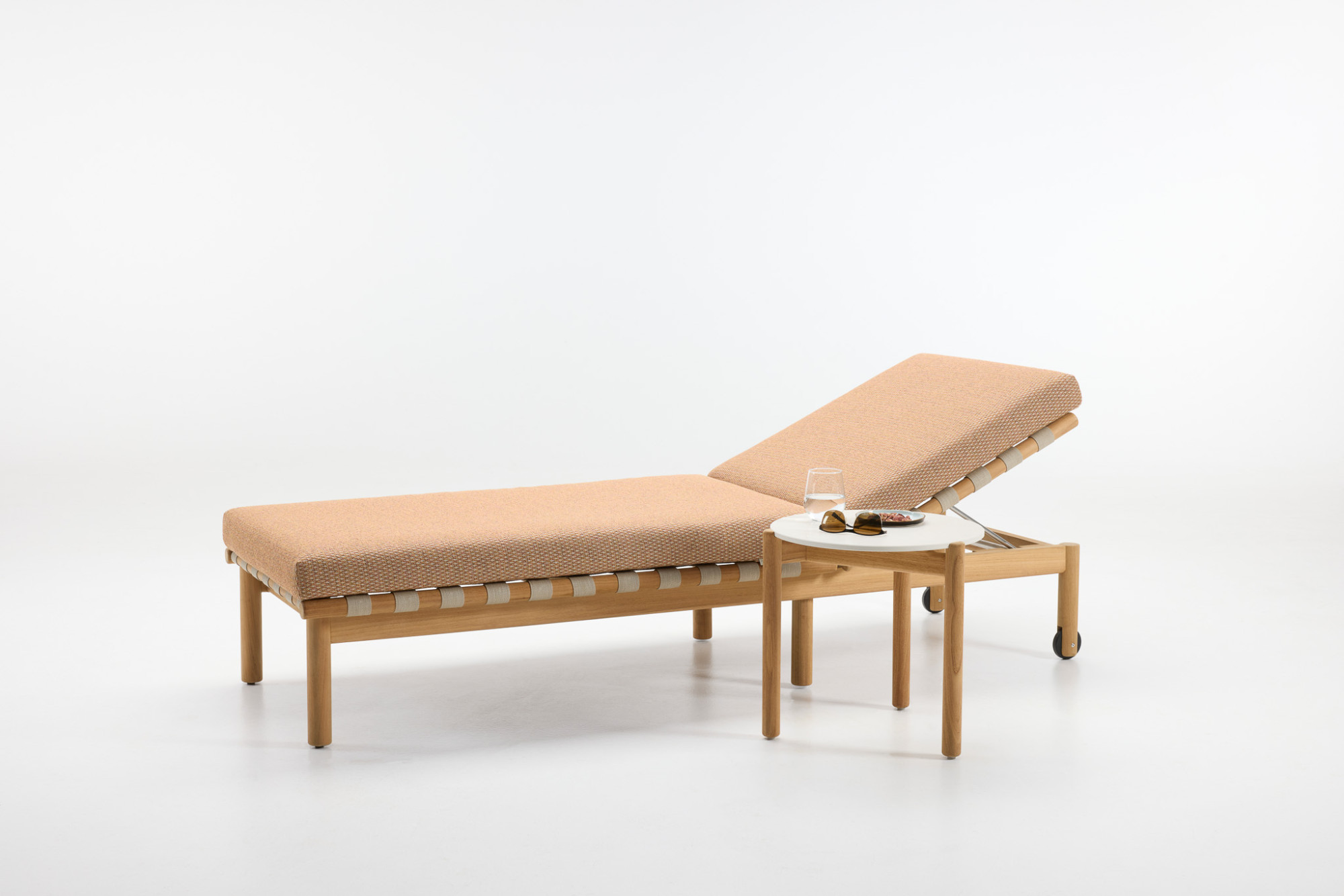 Chaise longue Oda en tissu couleur sienna avec cadre en teck, associée à la table basse Oda avec un plateau rond blanc, ornée de lunettes de soleil, d’un verre d’eau et d’une petite assiette, sur un fond blanc.