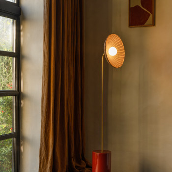 Arvin floor lamp with a curved brass stem, a handmade ceramic base in maroon, and a woven rattan shade, placed in a softly lit interior with earthy tones.